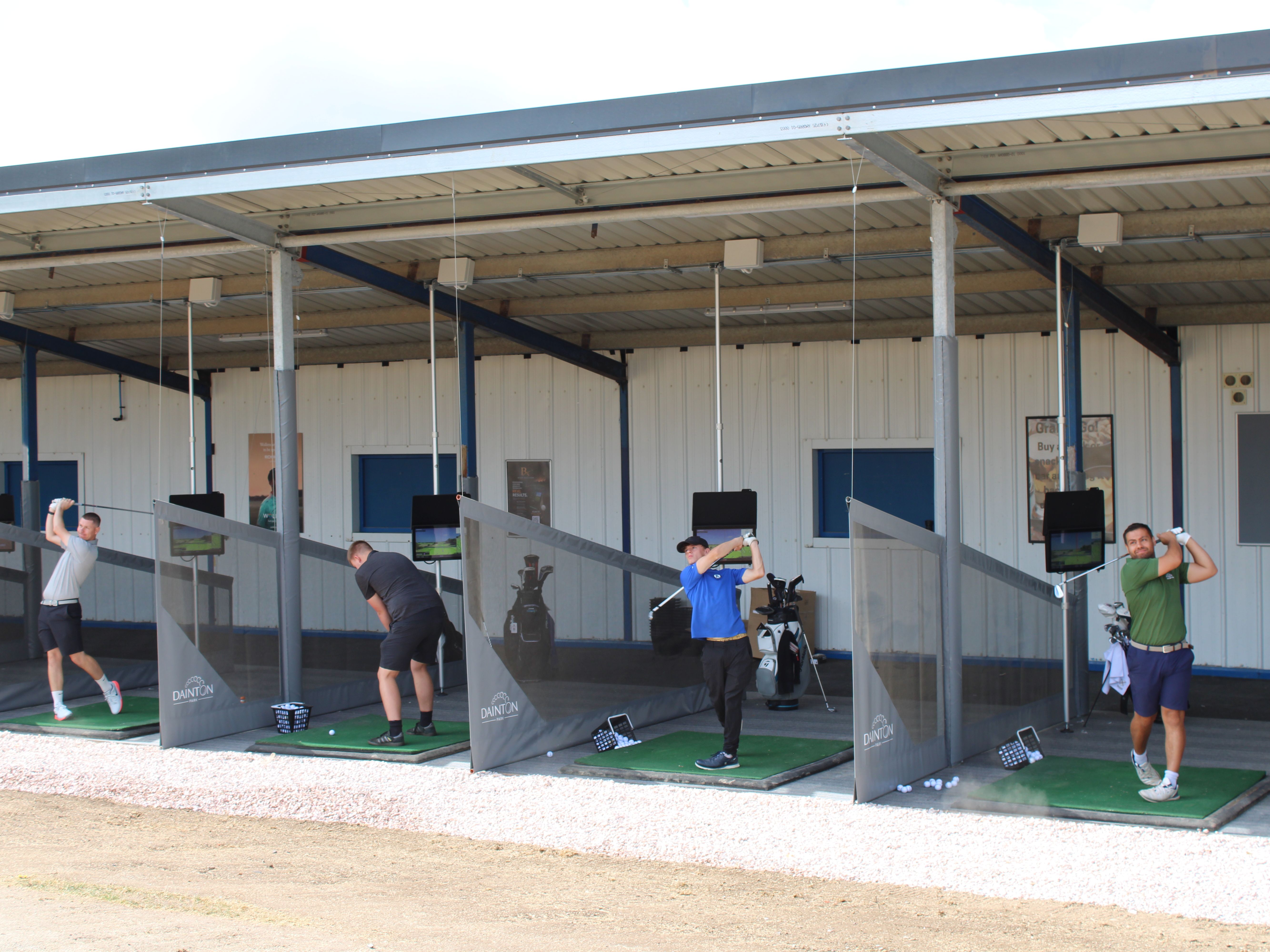 Golfers practising swings in covered driving range bays with netted dividers and launch monitors