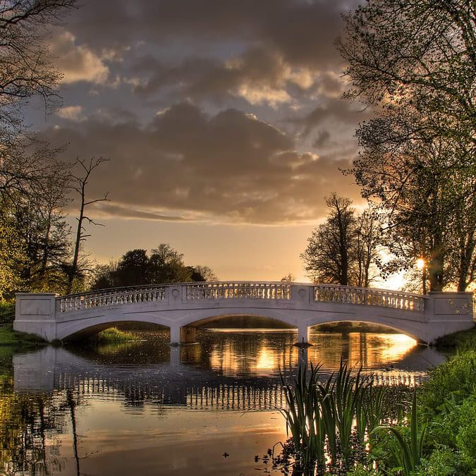 A stone bridge spans a calm pond surrounded by lush greenery and tall trees, reflecting the golden light of a sunset beneath a cloudy sky.