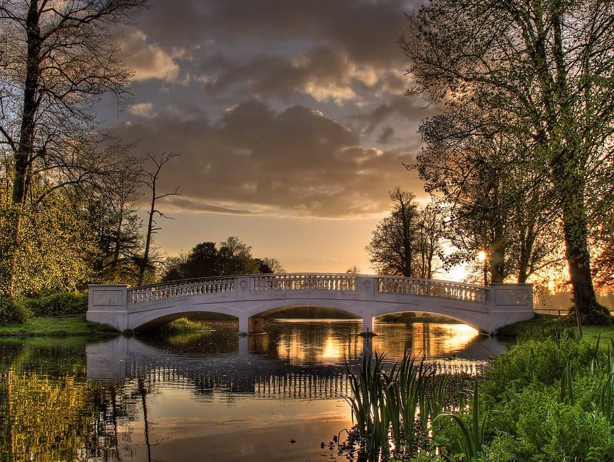 A stone bridge spans a calm pond surrounded by lush greenery and tall trees, reflecting the golden light of a sunset beneath a cloudy sky.