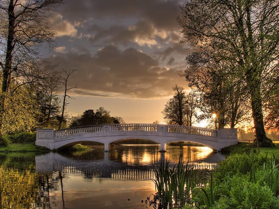 A stone bridge spans a calm pond surrounded by lush greenery and tall trees, reflecting the golden light of a sunset beneath a cloudy sky.
