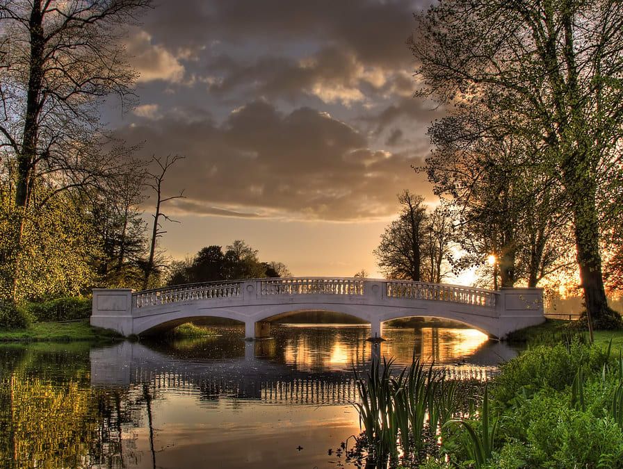 A stone bridge spans a calm pond surrounded by lush greenery and tall trees, reflecting the golden light of a sunset beneath a cloudy sky.