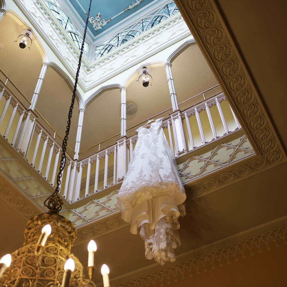 A wedding dress with intricate lace detail hangs over a balcony railing in a grand room with a large chandelier, ornate moldings, and a skylight above.