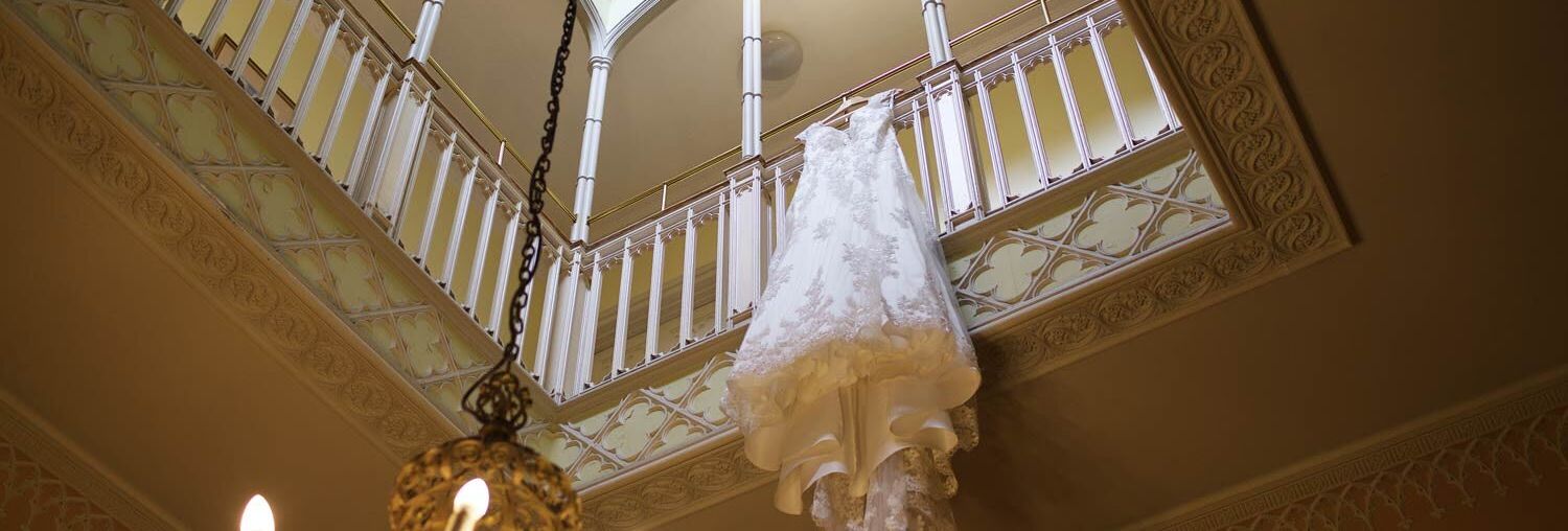 A wedding dress with intricate lace detail hangs over a balcony railing in a grand room with a large chandelier, ornate moldings, and a skylight above.