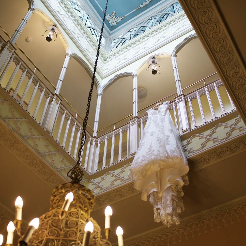 A wedding dress with intricate lace detail hangs over a balcony railing in a grand room with a large chandelier, ornate moldings, and a skylight above.