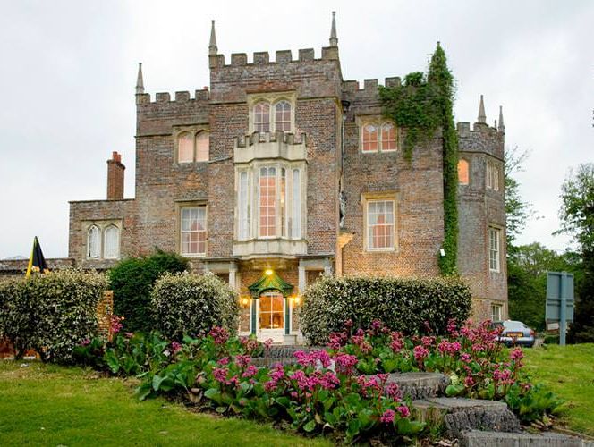 A historic brick building with a castle-like facade, featuring tall windows and ivy climbing its walls, sits behind a manicured garden with pink flowers.