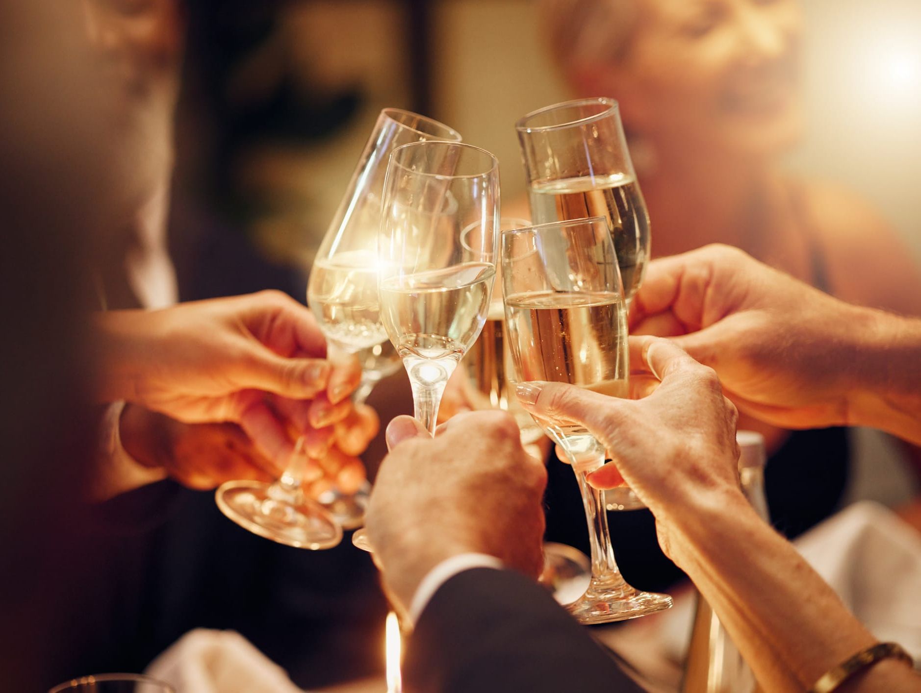 A group of people toasting with champagne glasses, celebrating in a warm, softly lit atmosphere, with hands raised in a celebratory gesture.
