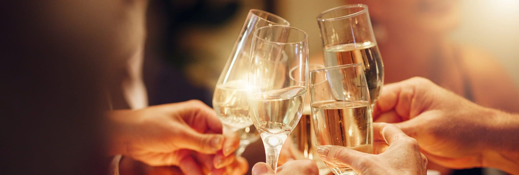 A group of people toasting with champagne glasses, celebrating in a warm, softly lit atmosphere, with hands raised in a celebratory gesture.