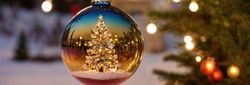 A reflective Christmas ornament hanging from a pine branch, showcasing a festive tree with colorful lights and ornaments, set against a snowy, softly lit background.