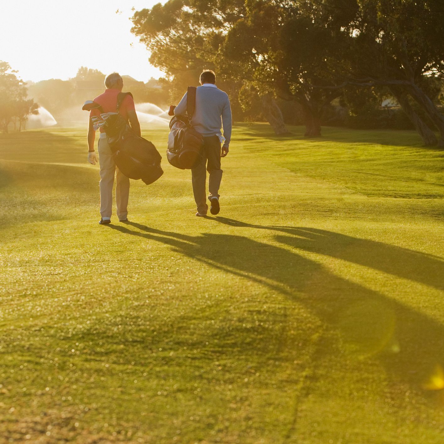 Two golfers walking across a sunlit fairway carrying their golf bags, with long shadows cast on the vibrant green grass and trees in the background.
