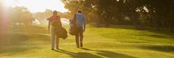 Two golfers walking across a sunlit fairway carrying their golf bags, with long shadows cast on the vibrant green grass and trees in the background.