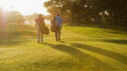 Two golfers walking across a sunlit fairway carrying their golf bags, with long shadows cast on the vibrant green grass and trees in the background.