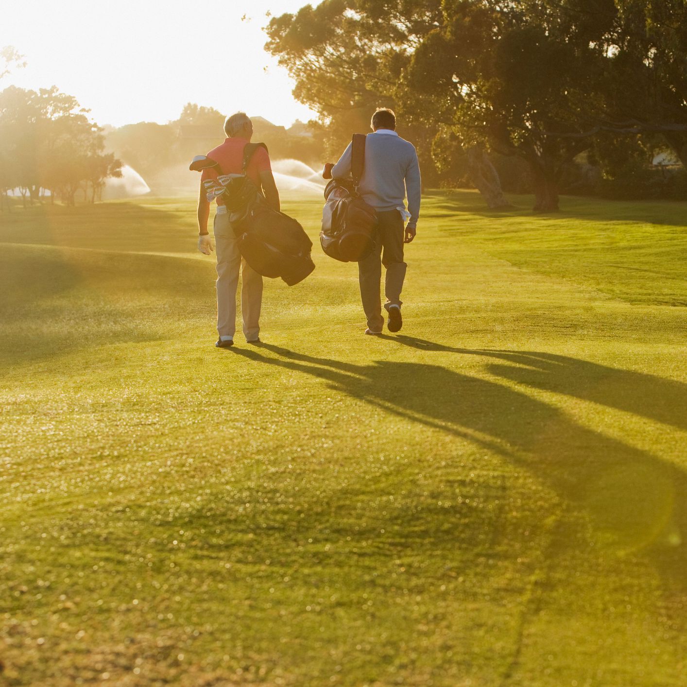 Two golfers walking across a sunlit fairway carrying their golf bags, with long shadows cast on the vibrant green grass and trees in the background.