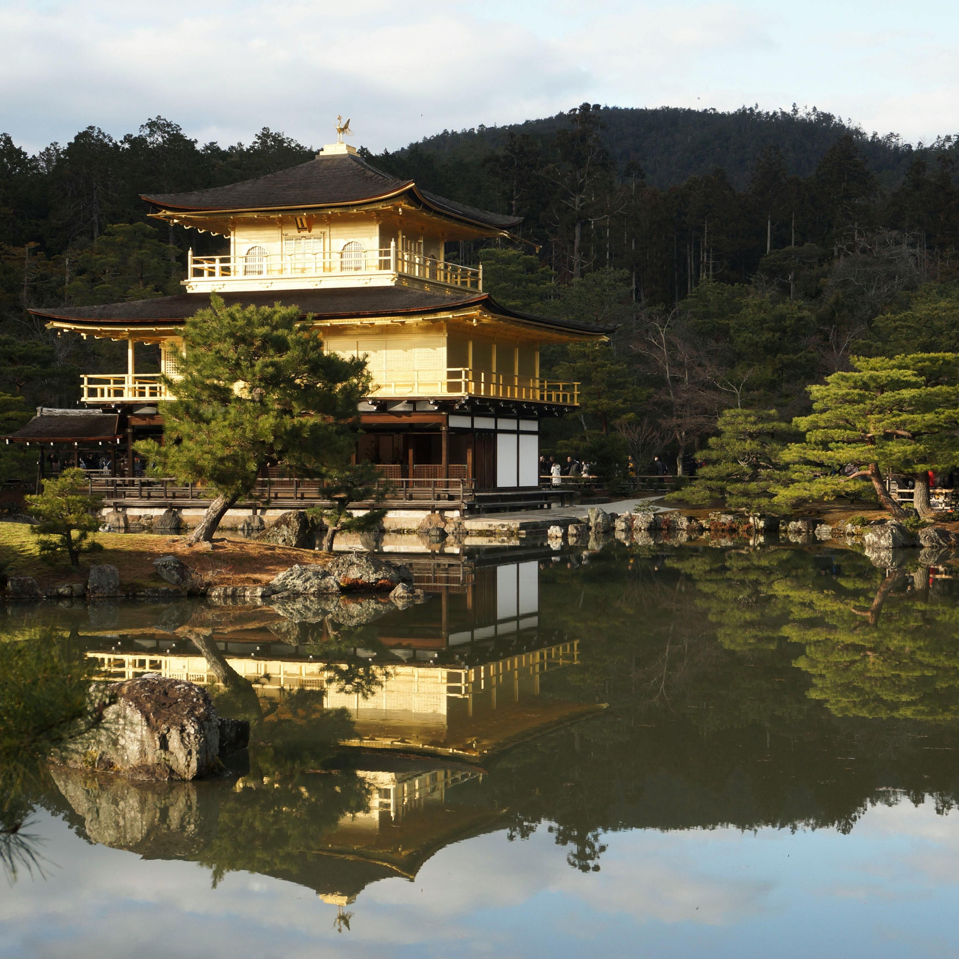 The Golden Pavilion (Kinkaku-ji), a traditional Japanese temple covered in gold leaf, reflected in a tranquil pond surrounded by lush greenery and forested hills.