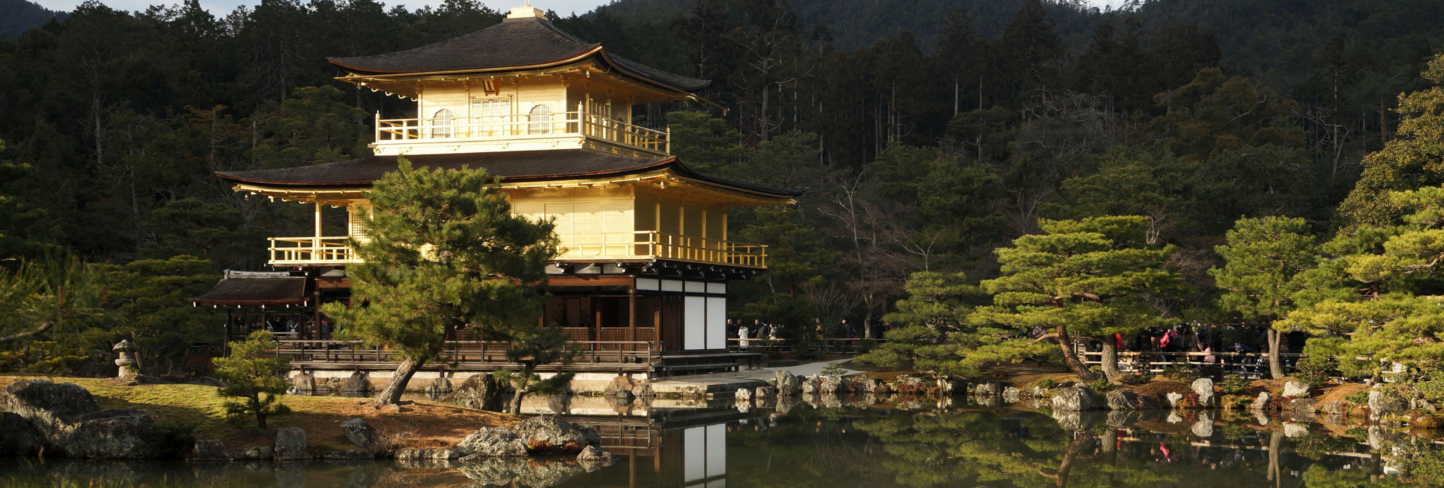 The Golden Pavilion (Kinkaku-ji), a traditional Japanese temple covered in gold leaf, reflected in a tranquil pond surrounded by lush greenery and forested hills.
