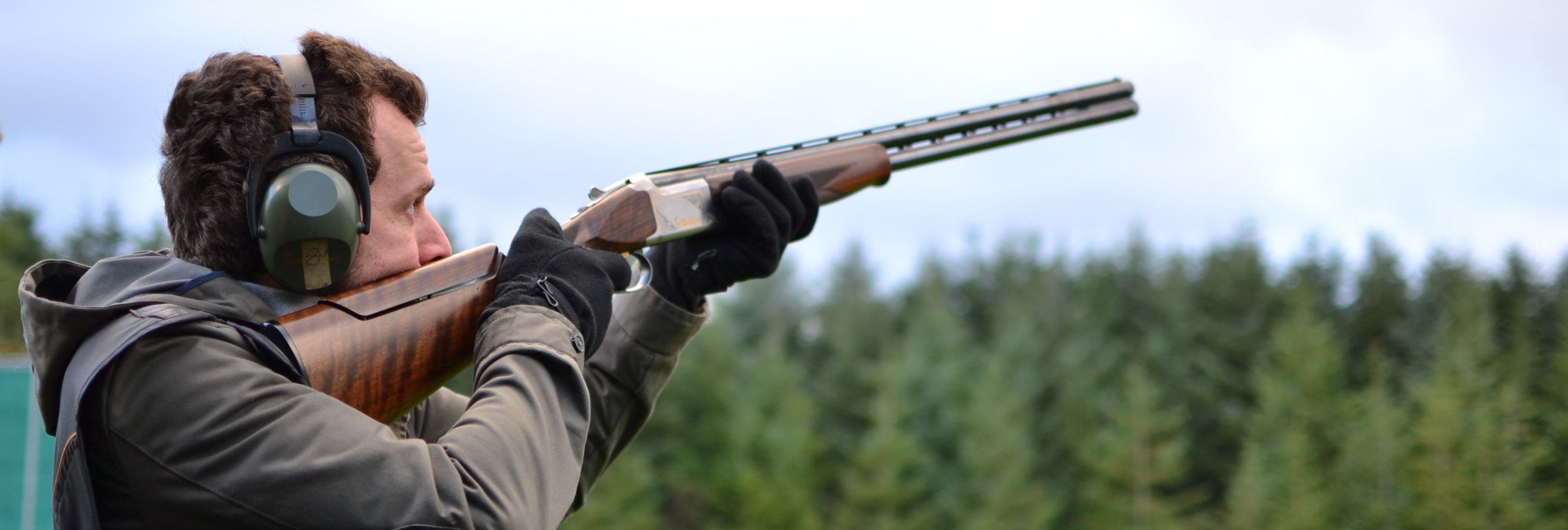 A man aiming a shotgun during target practice outdoors, wearing ear protection and gloves, with a blurred backdrop of green trees and a cloudy sky.