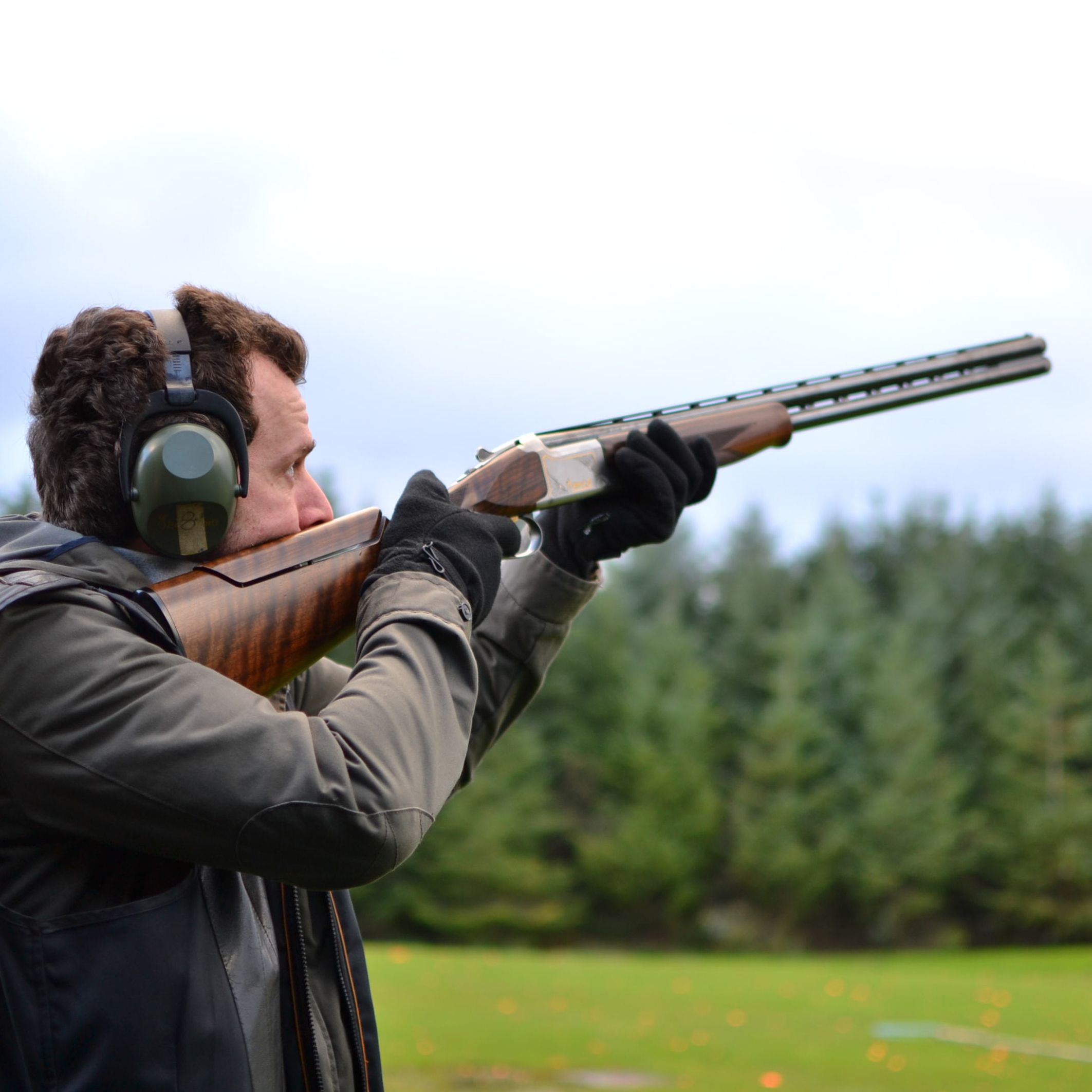 A man aiming a shotgun during target practice outdoors, wearing ear protection and gloves, with a blurred backdrop of green trees and a cloudy sky.