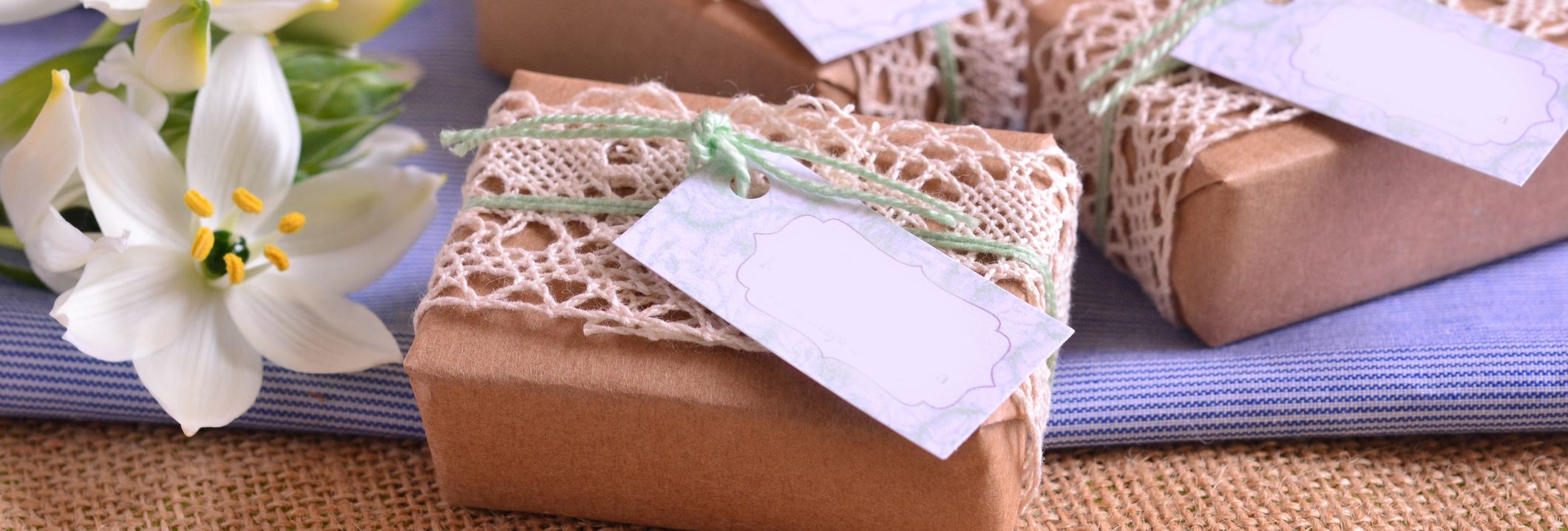 Handcrafted gift boxes wrapped in brown paper and lace, tied with green string and blank tags, arranged on a table with white lilies as decoration.