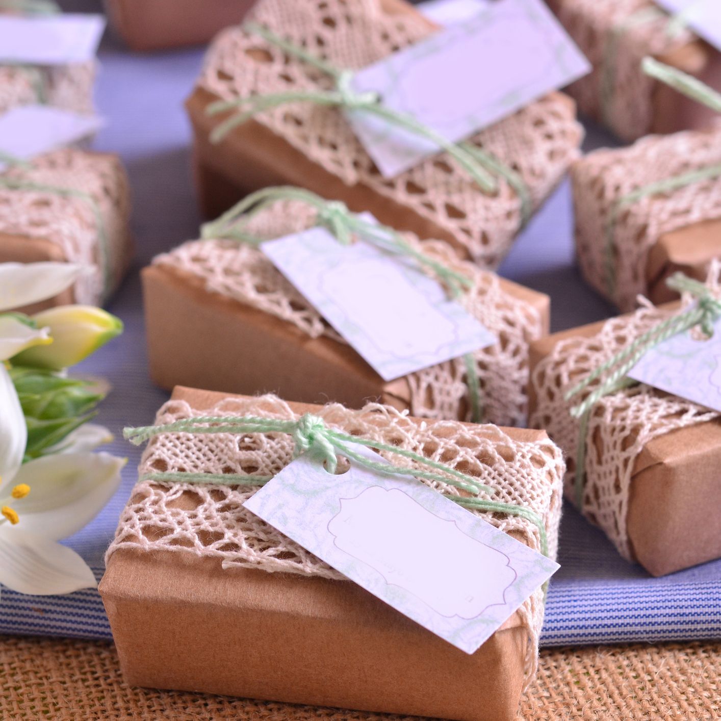 Handcrafted gift boxes wrapped in brown paper and lace, tied with green string and blank tags, arranged on a table with white lilies as decoration.