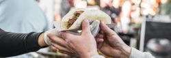 A close-up of hands exchanging a freshly made taco in a paper holder at an outdoor food market, with a blurred background of people and colorful stalls.