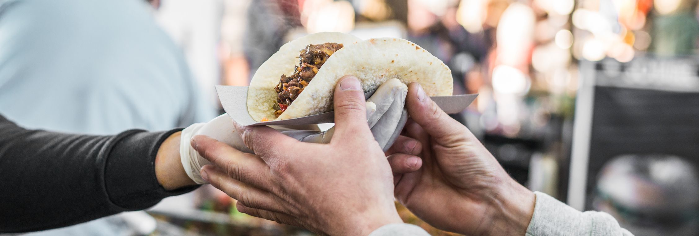 A close-up of hands exchanging a freshly made taco in a paper holder at an outdoor food market, with a blurred background of people and colorful stalls.