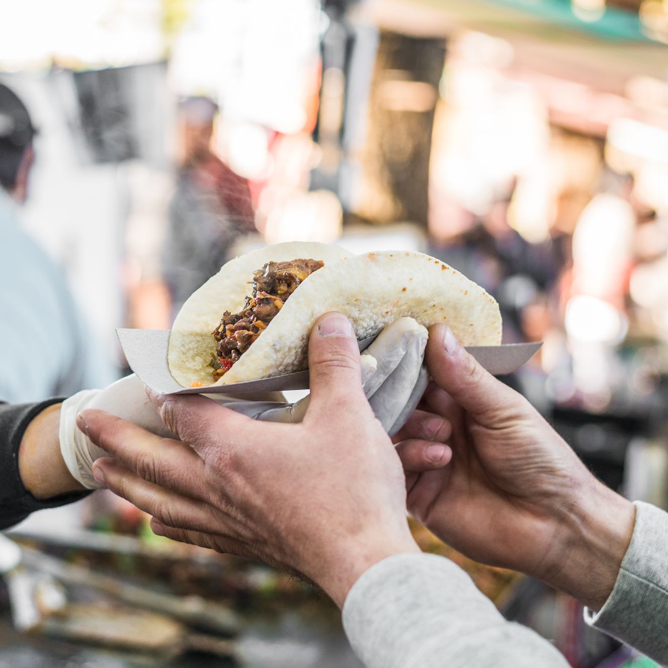 A close-up of hands exchanging a freshly made taco in a paper holder at an outdoor food market, with a blurred background of people and colorful stalls.