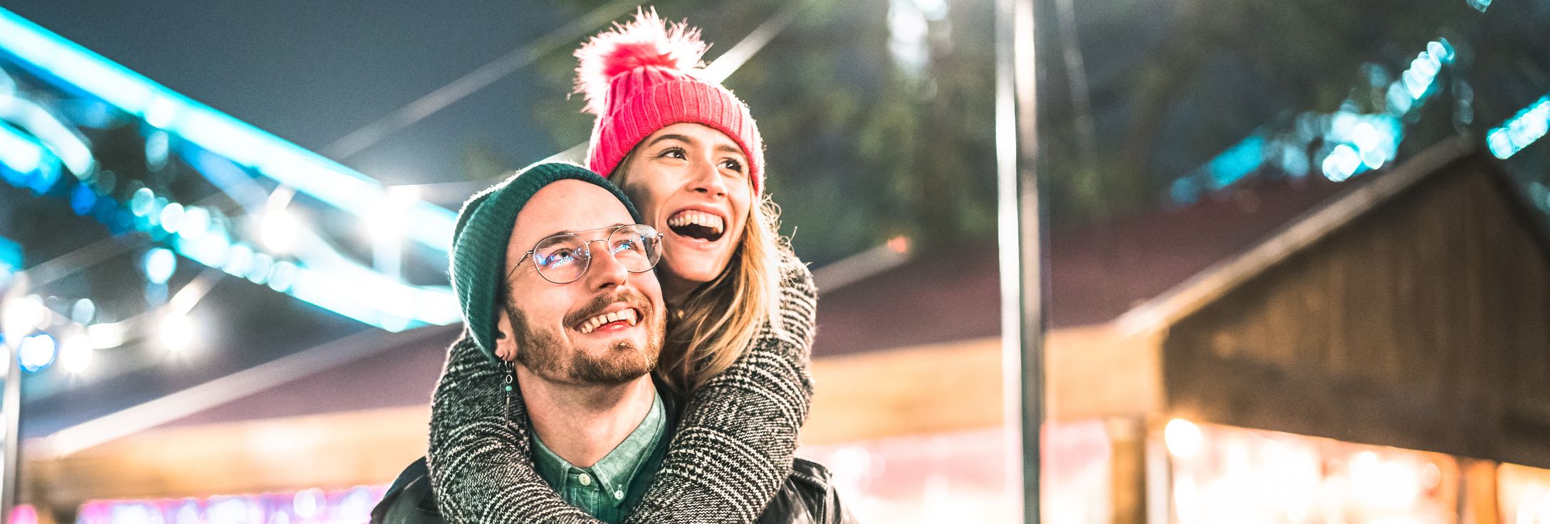 A joyful couple at a festive outdoor event, with the woman in a pink beanie playfully hugging the man from behind as they smile under colorful lights and decorations.