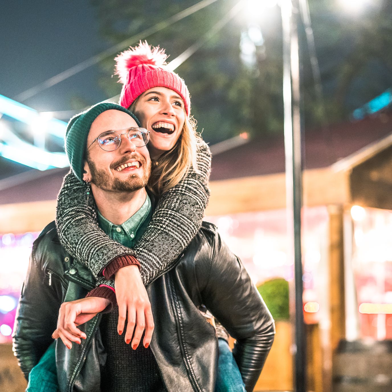 A joyful couple at a festive outdoor event, with the woman in a pink beanie playfully hugging the man from behind as they smile under colorful lights and decorations.