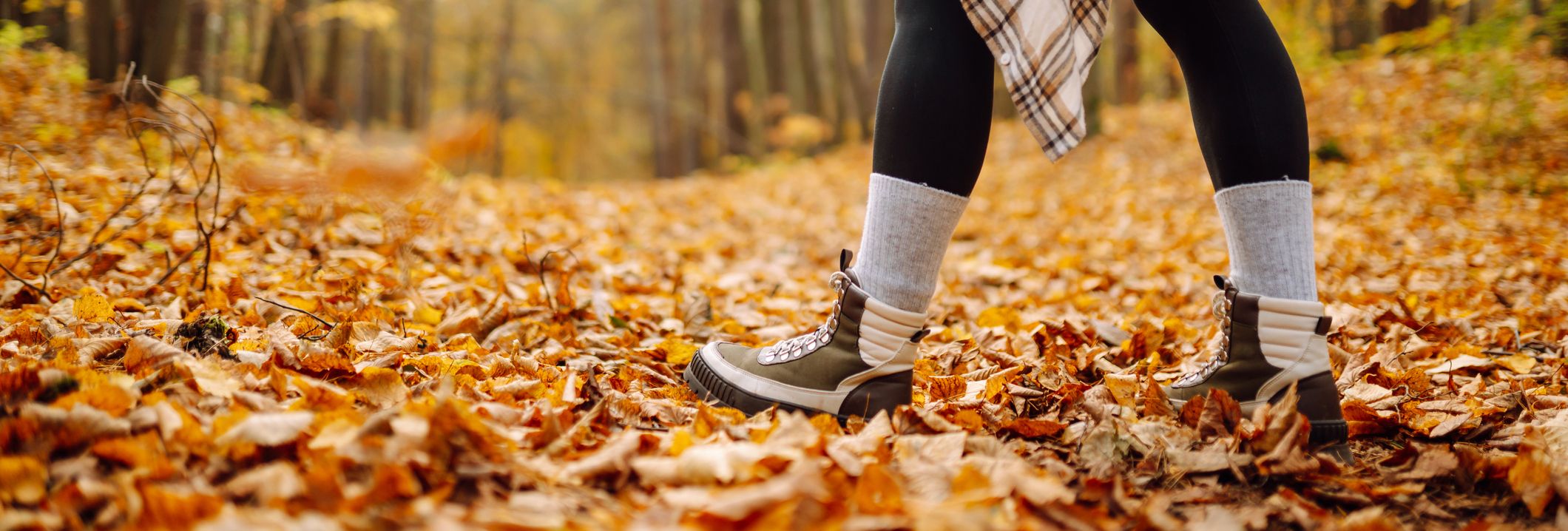 A person walking through an autumn forest, wearing boots, leggings, and a plaid shirt tied around their waist, with the ground covered in golden fallen leaves.