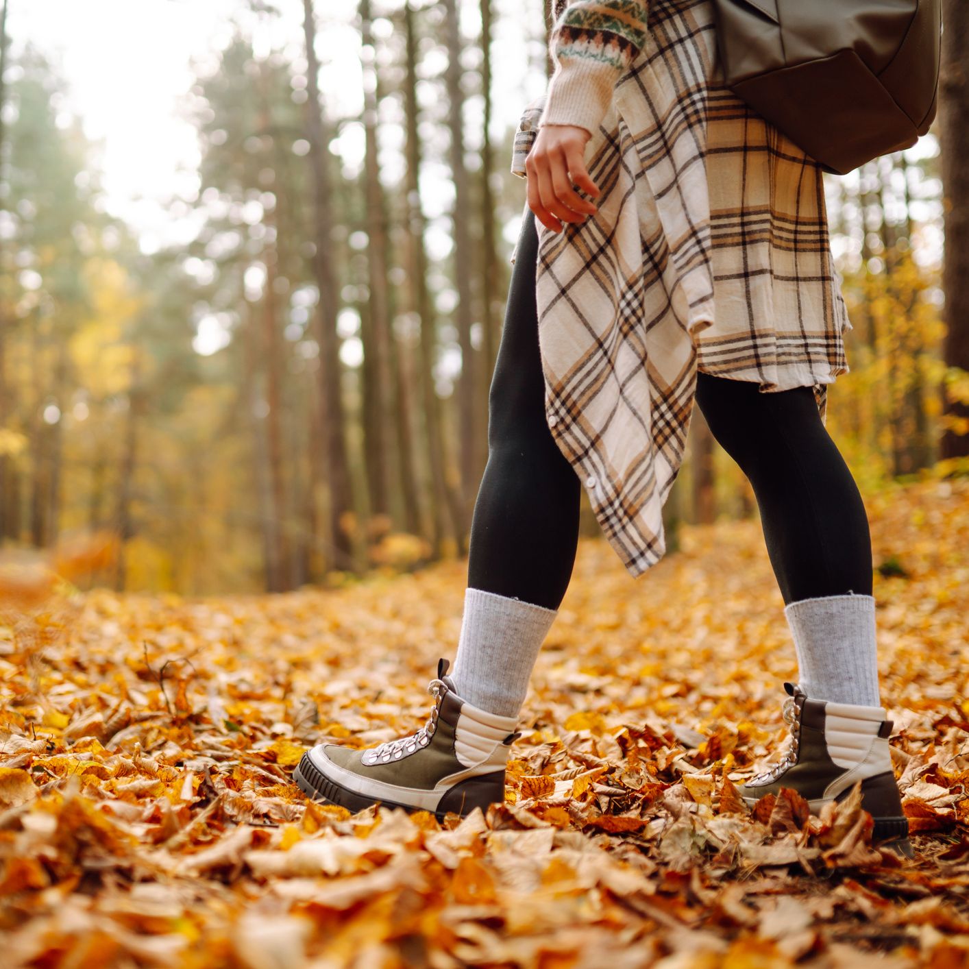 A person walking through an autumn forest, wearing boots, leggings, and a plaid shirt tied around their waist, with the ground covered in golden fallen leaves.