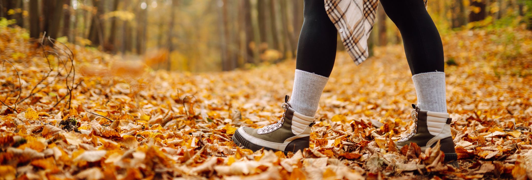 A person walking through an autumn forest, wearing boots, leggings, and a plaid shirt tied around their waist, with the ground covered in golden fallen leaves.