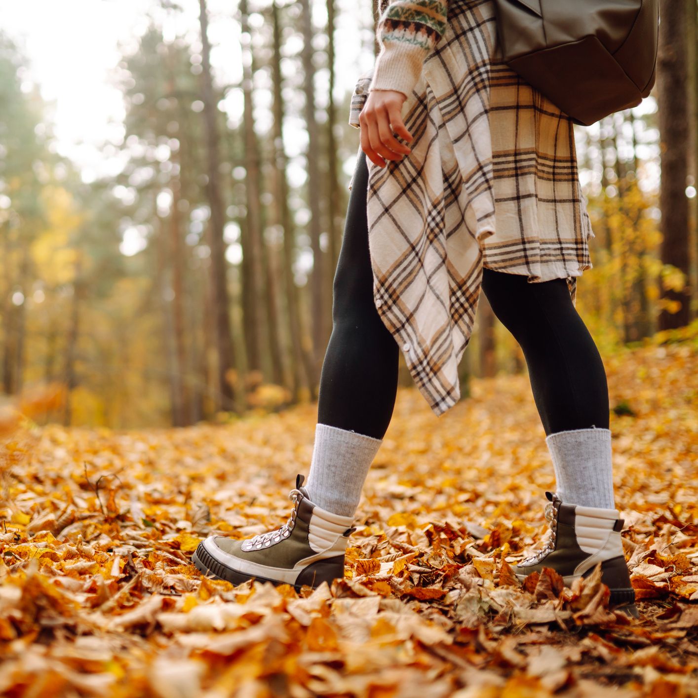 A person walking through an autumn forest, wearing boots, leggings, and a plaid shirt tied around their waist, with the ground covered in golden fallen leaves.