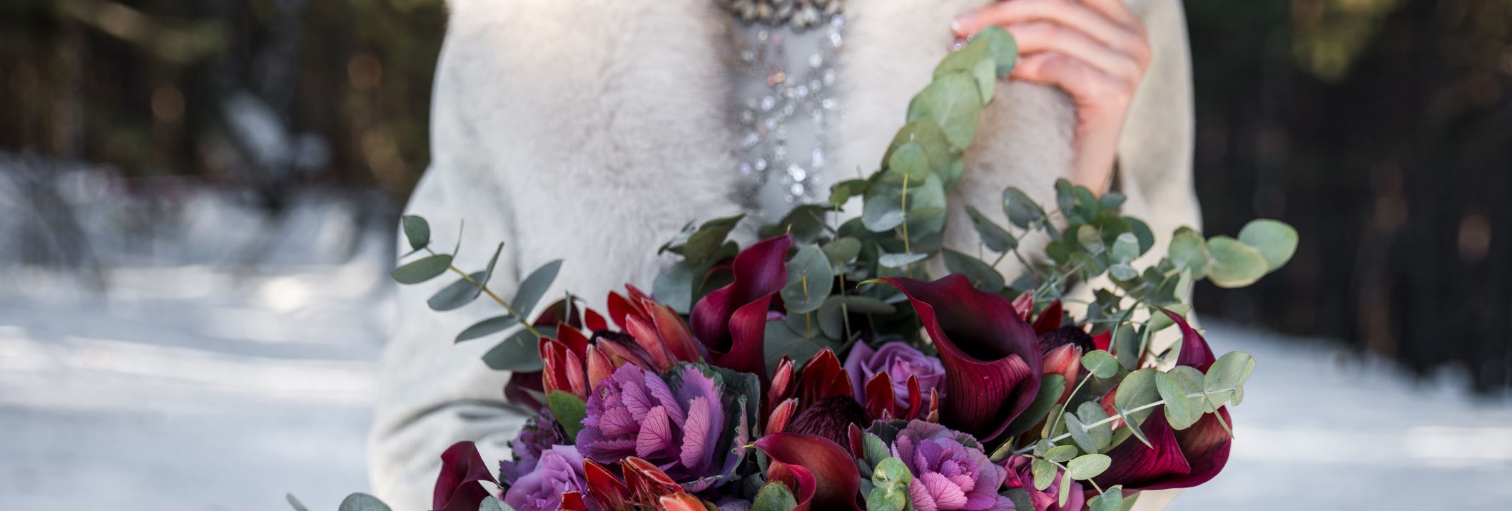 A person in a light coat holding a vibrant bouquet of red and purple flowers with eucalyptus leaves, set against a snowy outdoor background.