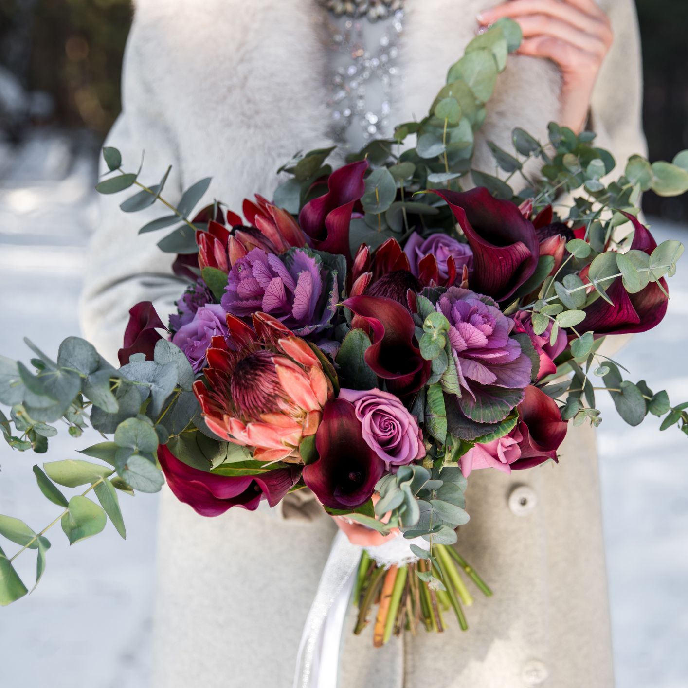 A person in a light coat holding a vibrant bouquet of red and purple flowers with eucalyptus leaves, set against a snowy outdoor background.