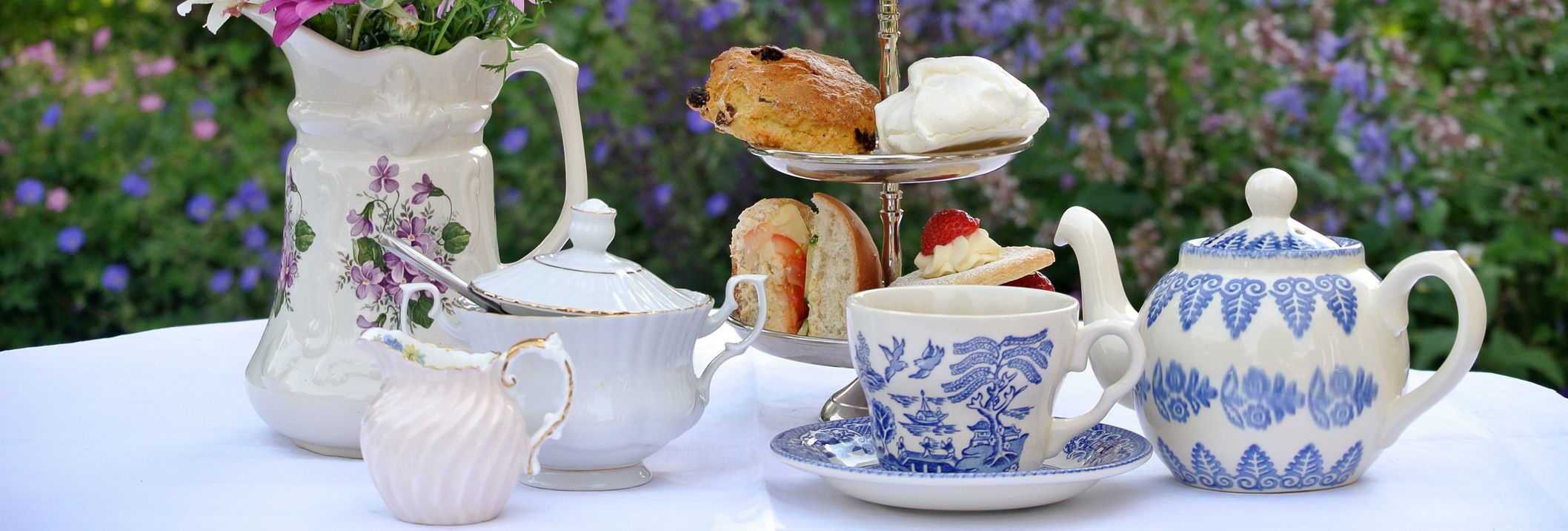 An elegant afternoon tea setup with a tiered tray of scones, sandwiches, and strawberries, surrounded by floral teapots, cups, and a vase of fresh flowers in a garden setting.