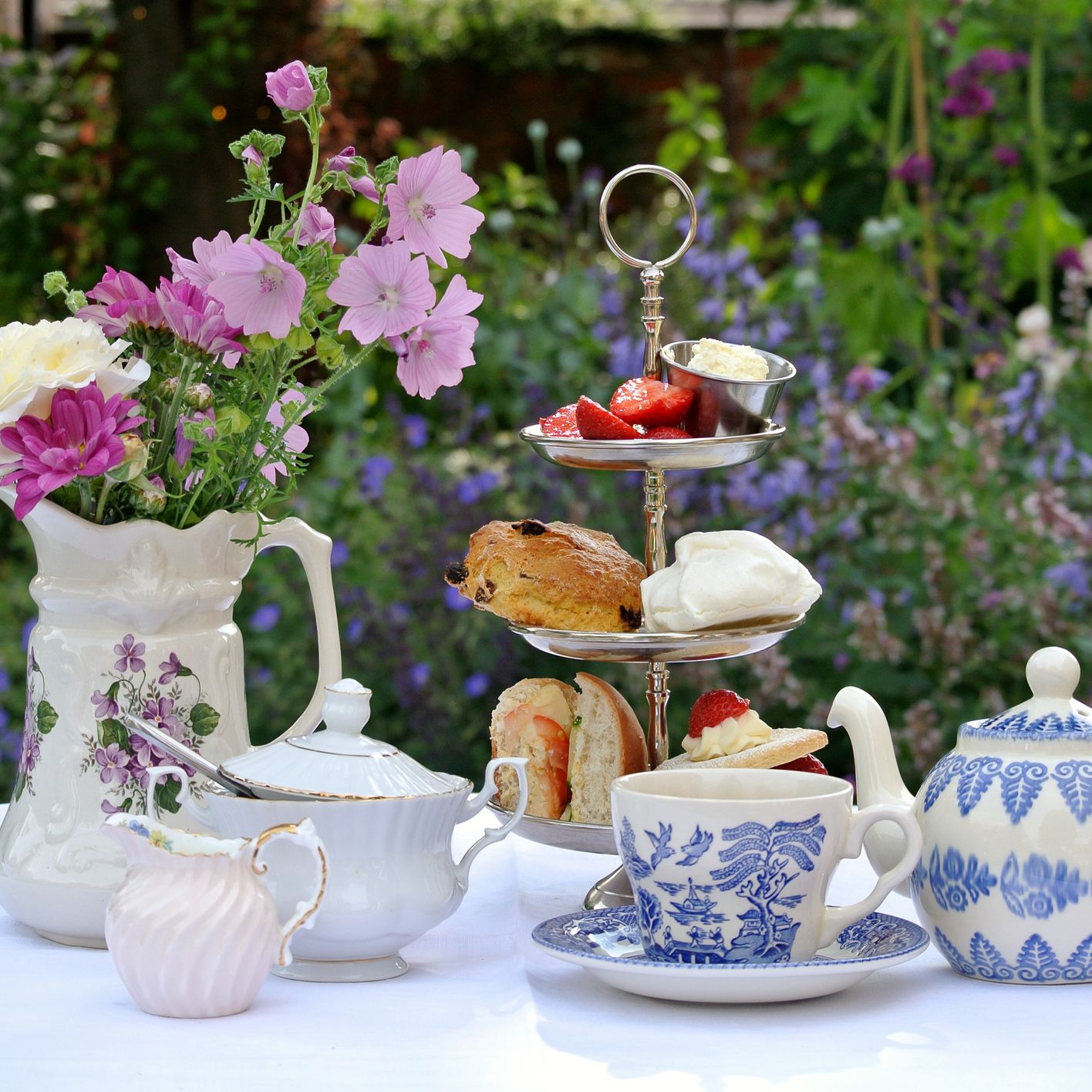 An elegant afternoon tea setup with a tiered tray of scones, sandwiches, and strawberries, surrounded by floral teapots, cups, and a vase of fresh flowers in a garden setting.