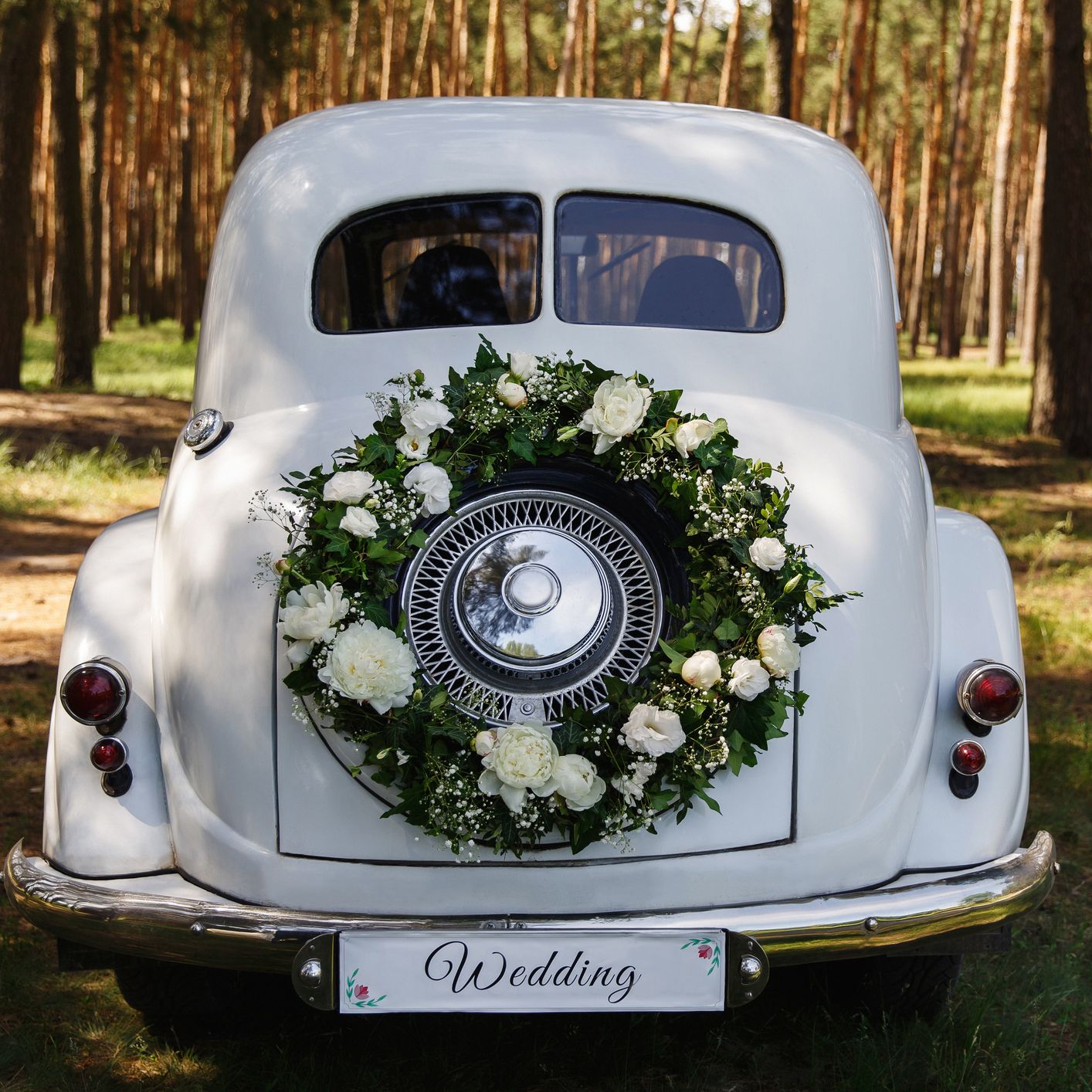 The rear of a vintage white car decorated with a wreath of white flowers and greenery, parked in a sunlit forest, with a 'Wedding' sign on the bumper.