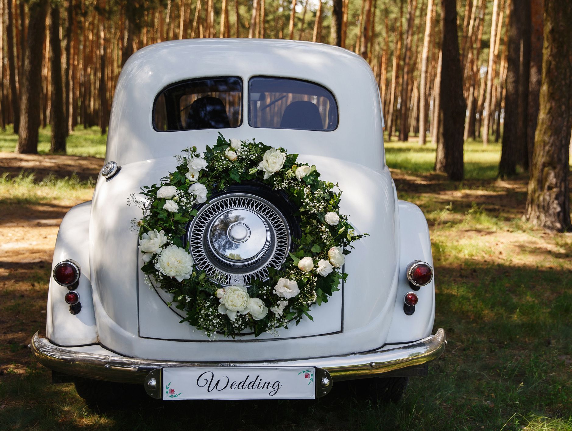 The rear of a vintage white car decorated with a wreath of white flowers and greenery, parked in a sunlit forest, with a 'Wedding' sign on the bumper.