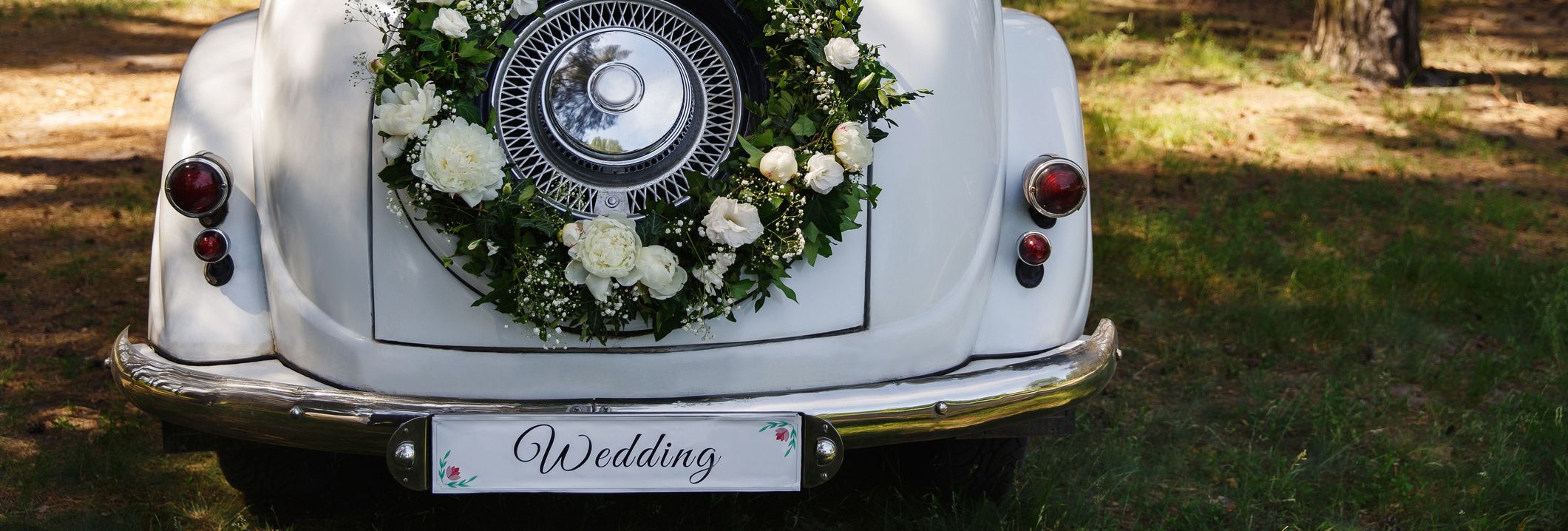 The rear of a vintage white car decorated with a wreath of white flowers and greenery, parked in a sunlit forest, with a 'Wedding' sign on the bumper.
