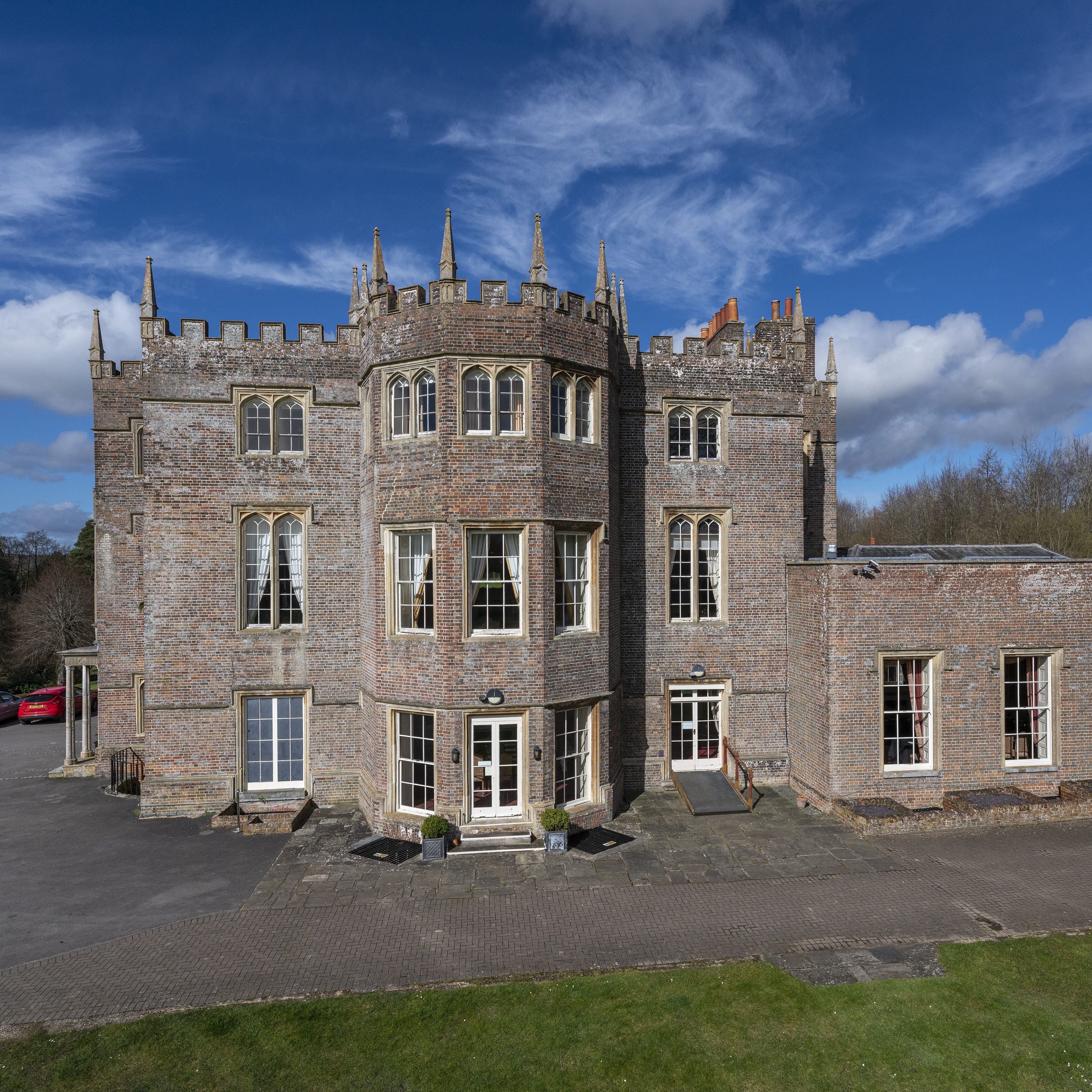 Donnington Grove with a castle-like design, featuring tall windows, battlements, and a central turret, set against a bright blue sky and surrounded by greenery.
