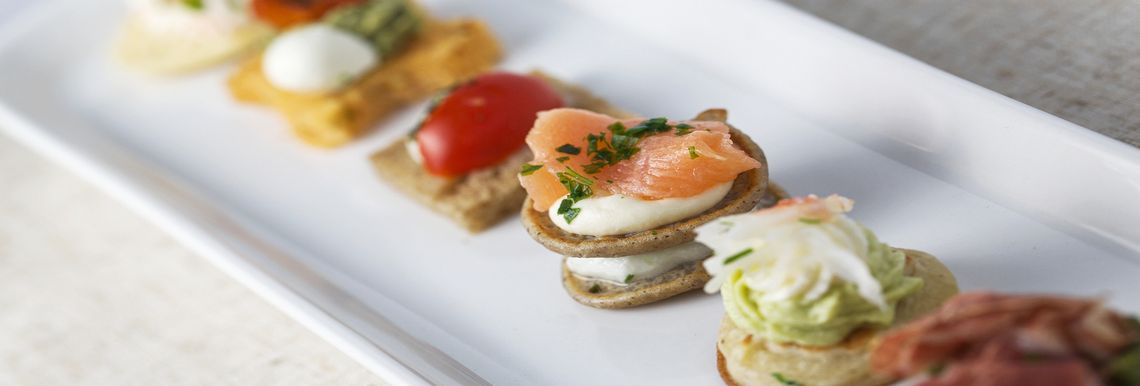 A close-up of an elegant platter showcasing assorted bite-sized appetizers, including smoked salmon, cherry tomatoes, and whipped toppings, arranged on a white rectangular plate.