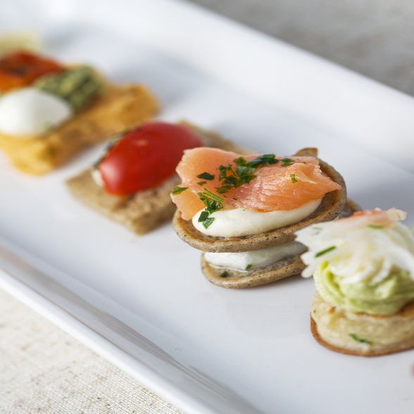 A close-up of an elegant platter showcasing assorted bite-sized appetizers, including smoked salmon, cherry tomatoes, and whipped toppings, arranged on a white rectangular plate.