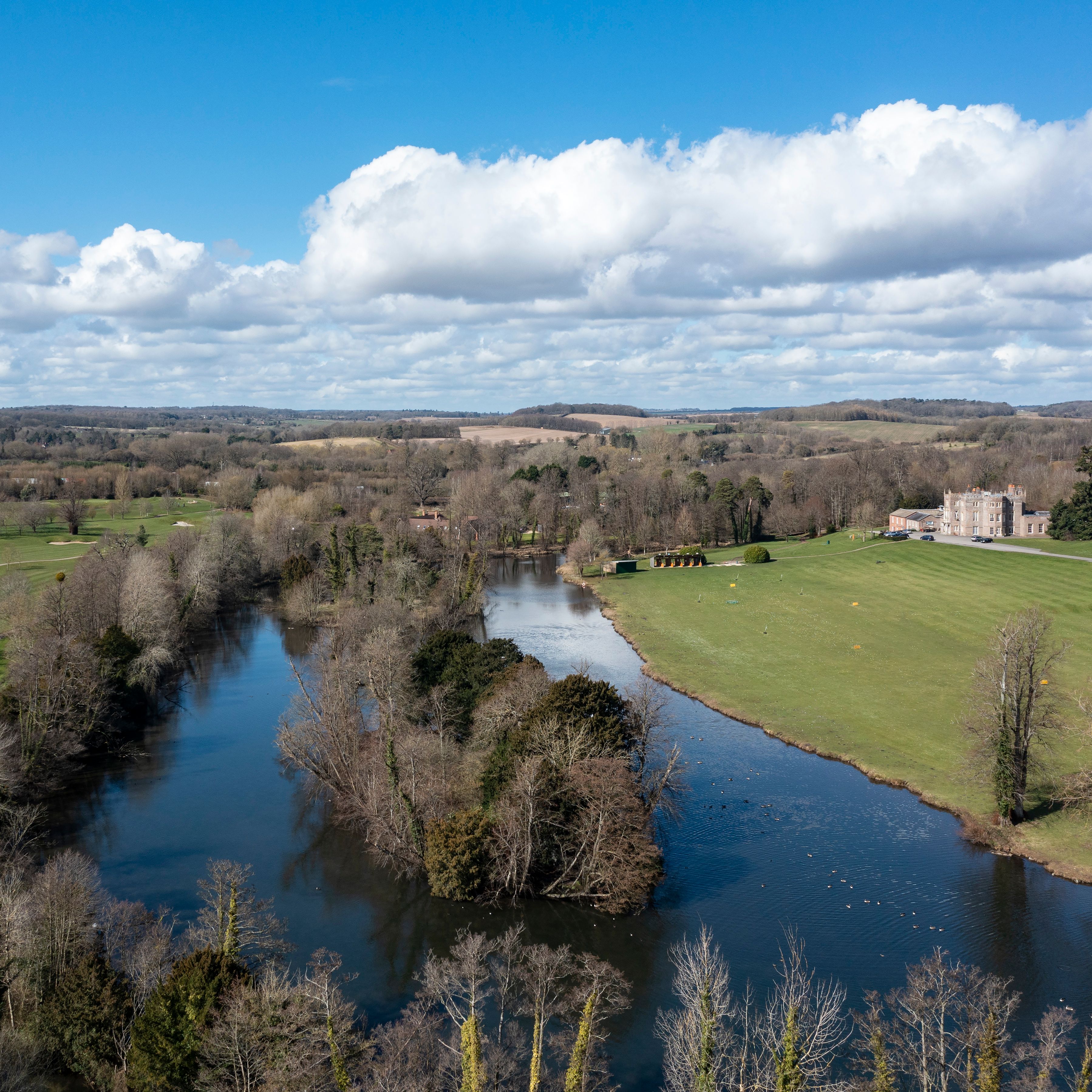 An aerial view of a countryside with a winding river, surrounding trees, grassy fields, and a historic manor in the distance under a bright blue sky with scattered clouds.