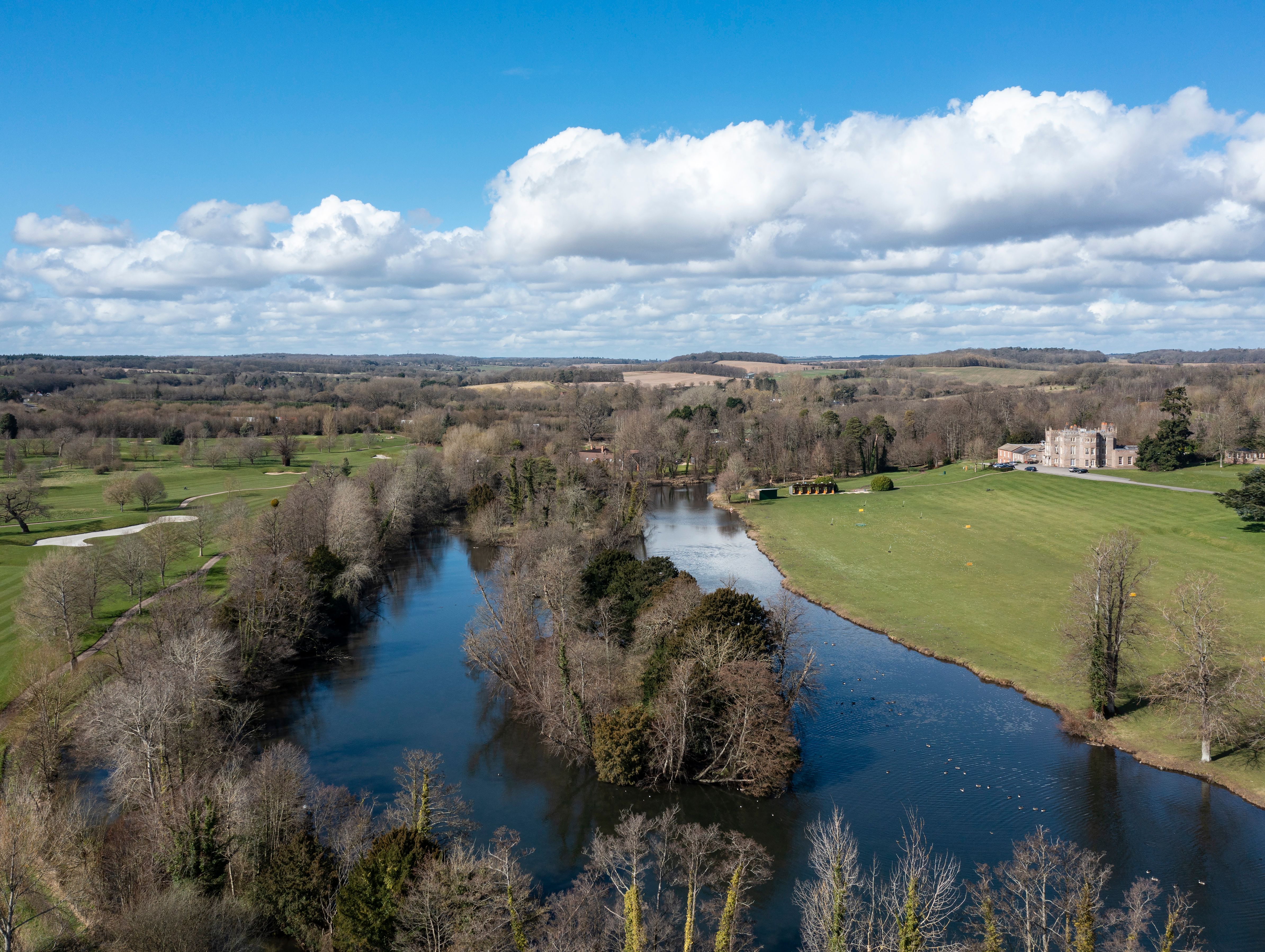An aerial view of a countryside with a winding river, surrounding trees, grassy fields, and a historic manor in the distance under a bright blue sky with scattered clouds.
