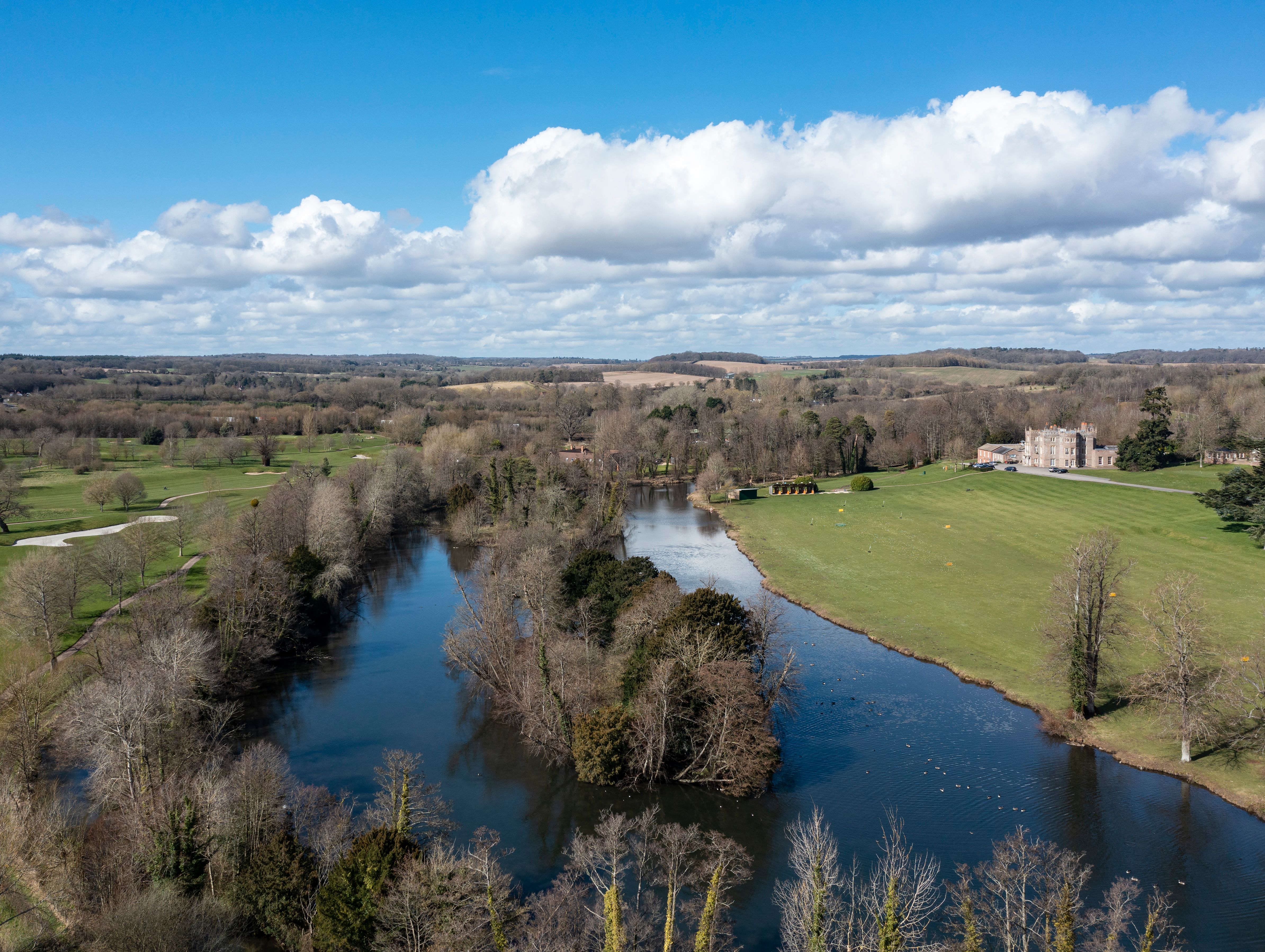 An aerial view of a countryside with a winding river, surrounding trees, grassy fields, and a historic manor in the distance under a bright blue sky with scattered clouds.