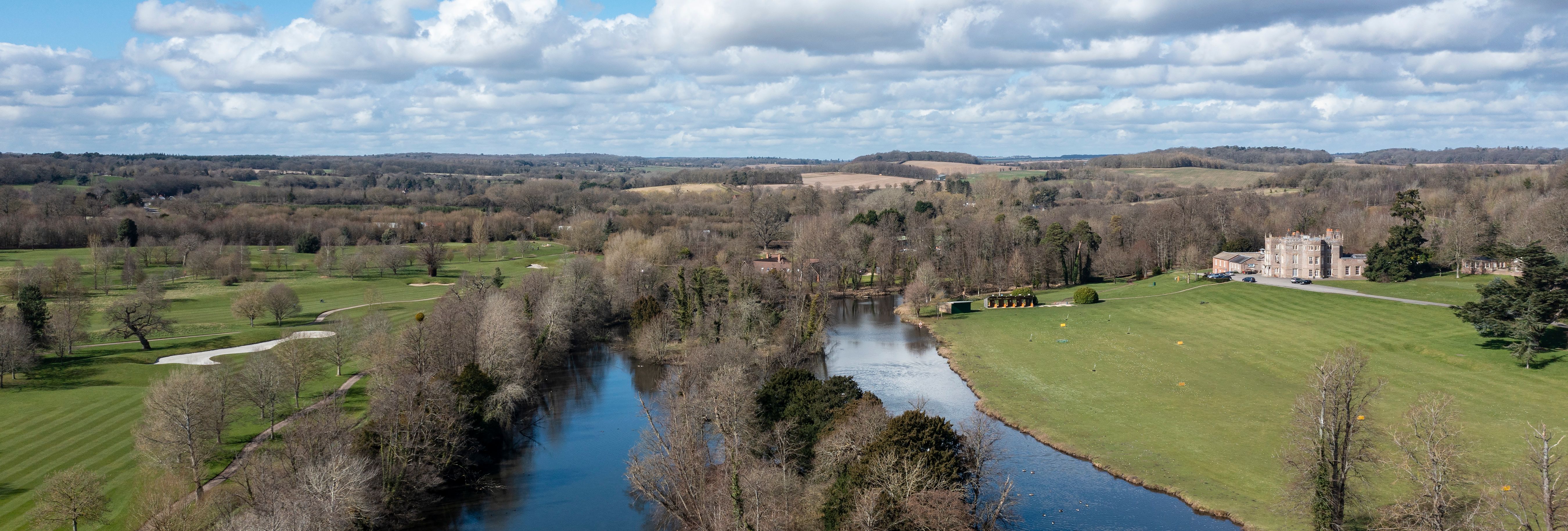 An aerial view of a countryside with a winding river, surrounding trees, grassy fields, and a historic manor in the distance under a bright blue sky with scattered clouds.