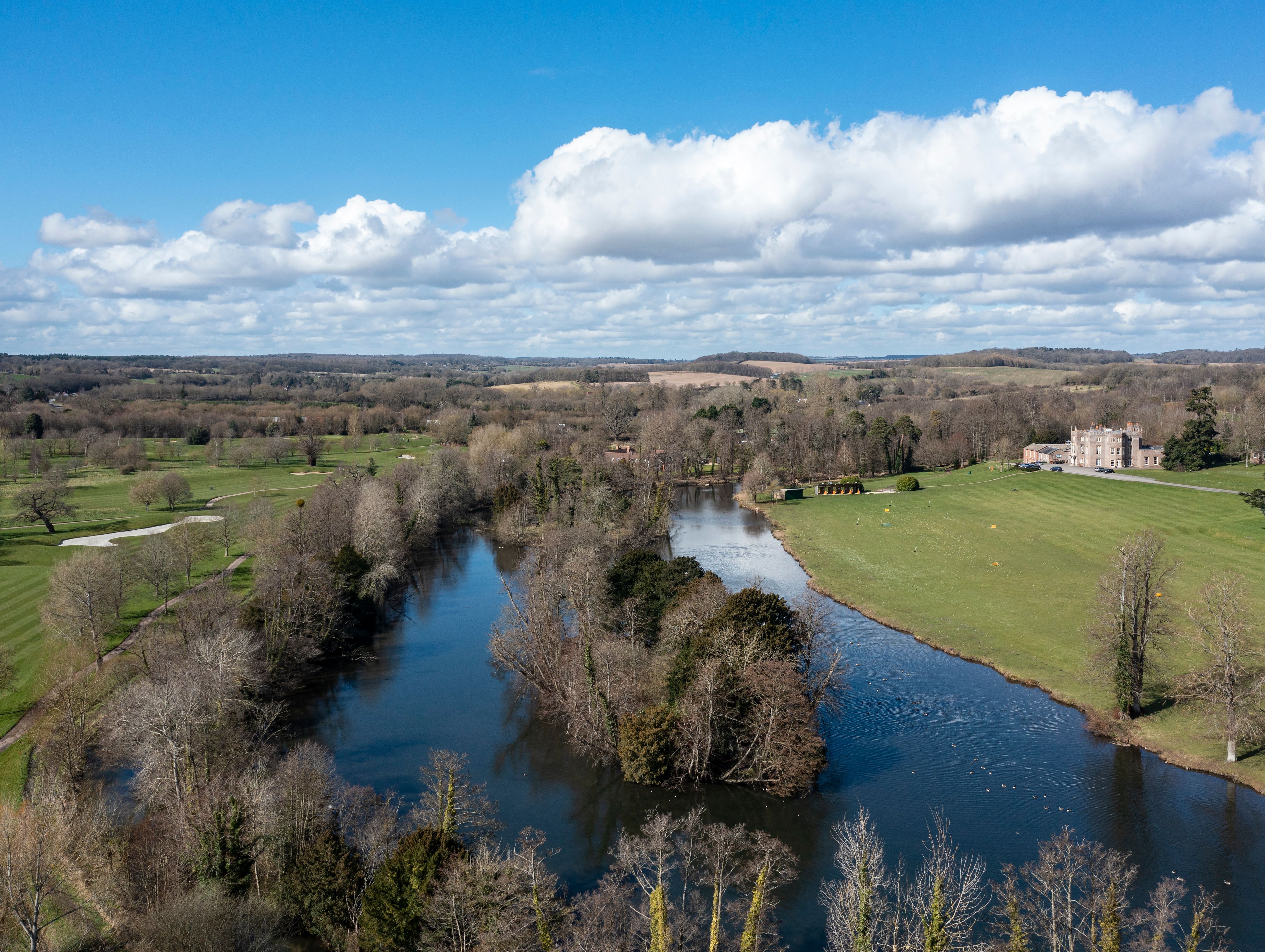 An aerial view of a countryside with a winding river, surrounding trees, grassy fields, and a historic manor in the distance under a bright blue sky with scattered clouds.
