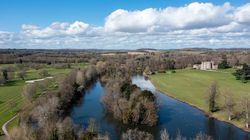 An aerial view of a countryside with a winding river, surrounding trees, grassy fields, and a historic manor in the distance under a bright blue sky with scattered clouds.