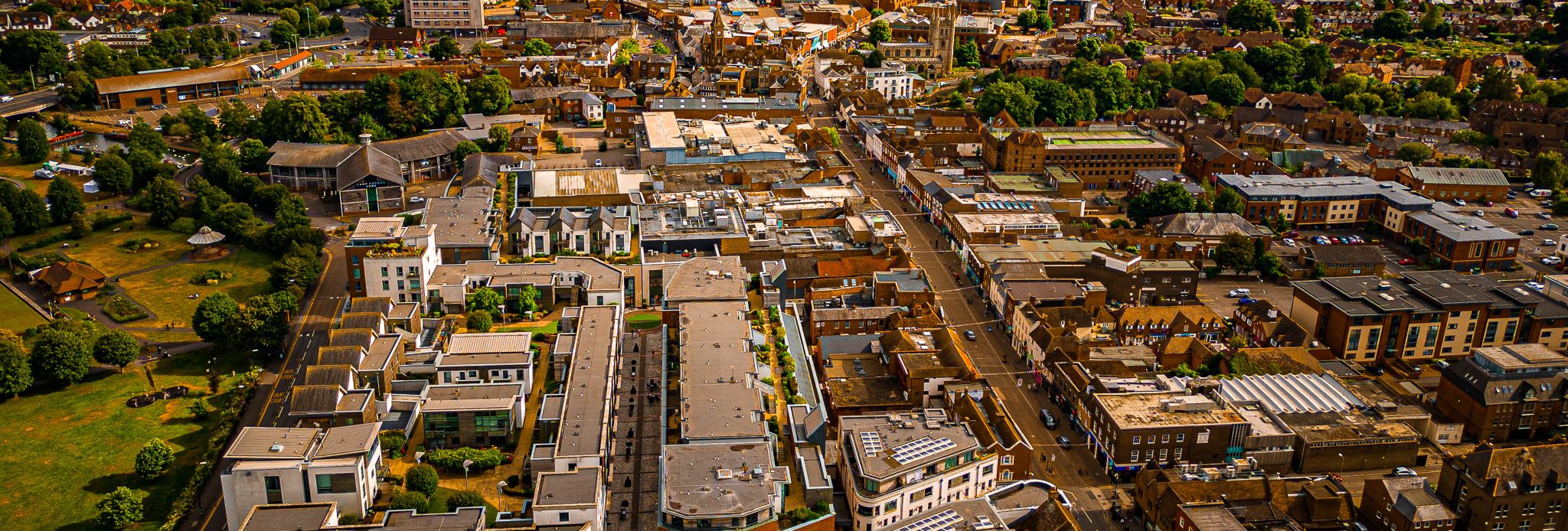 Aerial view of a town with modern buildings, tree-lined streets, and green parks under a partly cloudy sky, stretching into the distance.