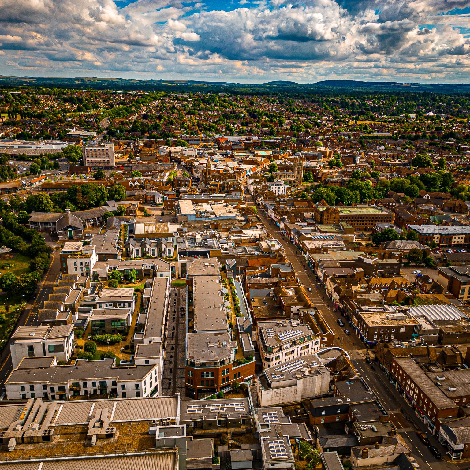 Aerial view of a town with modern buildings, tree-lined streets, and green parks under a partly cloudy sky, stretching into the distance.