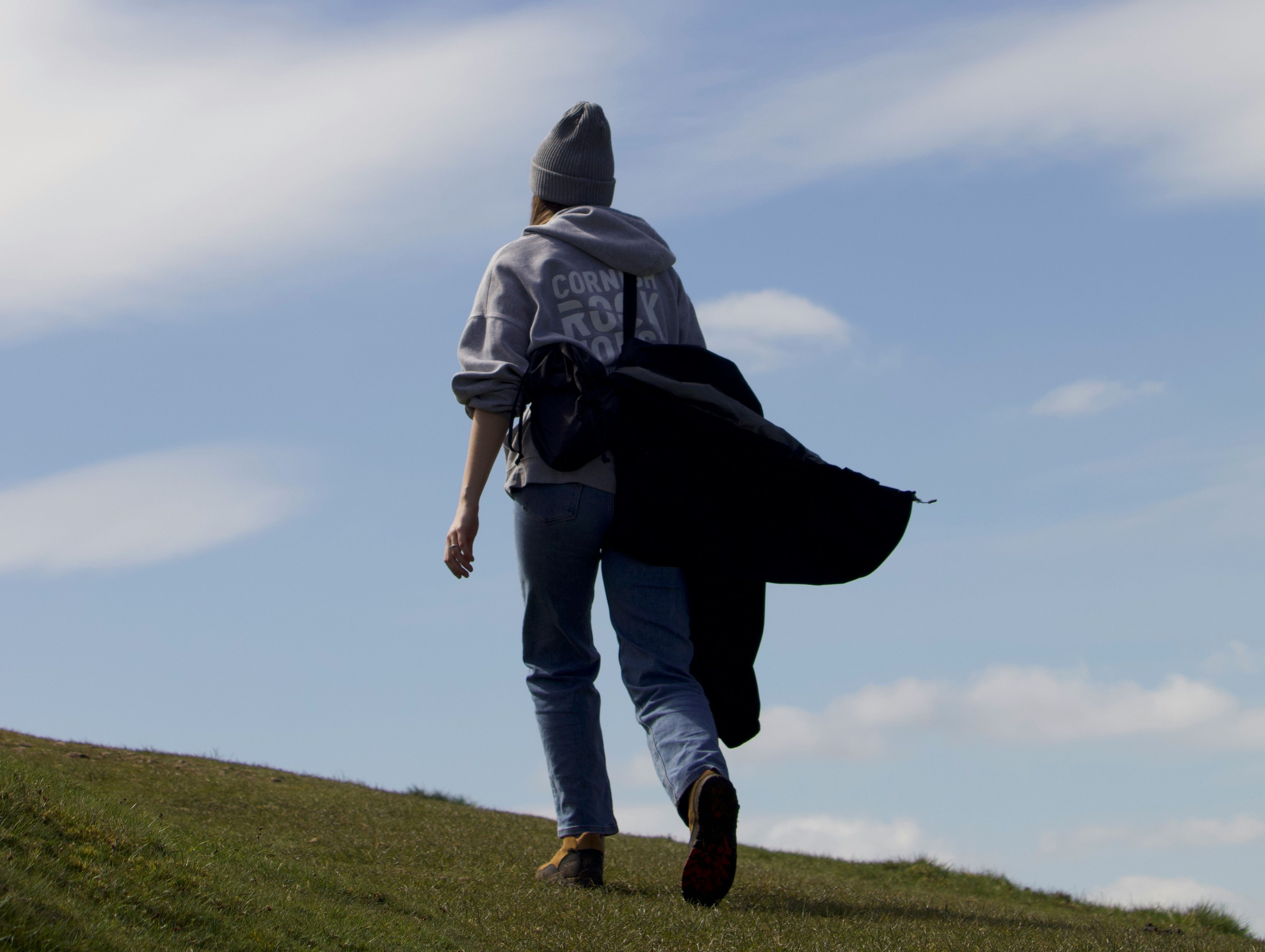 A person wearing a hoodie, jeans, and a beanie walks uphill on a grassy landscape.